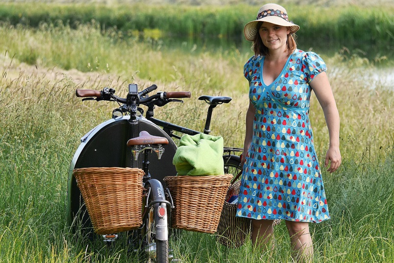 Ulrike trägt ein blaues Sommerkleid und steht neben dem Lastenrad im Kornfeld